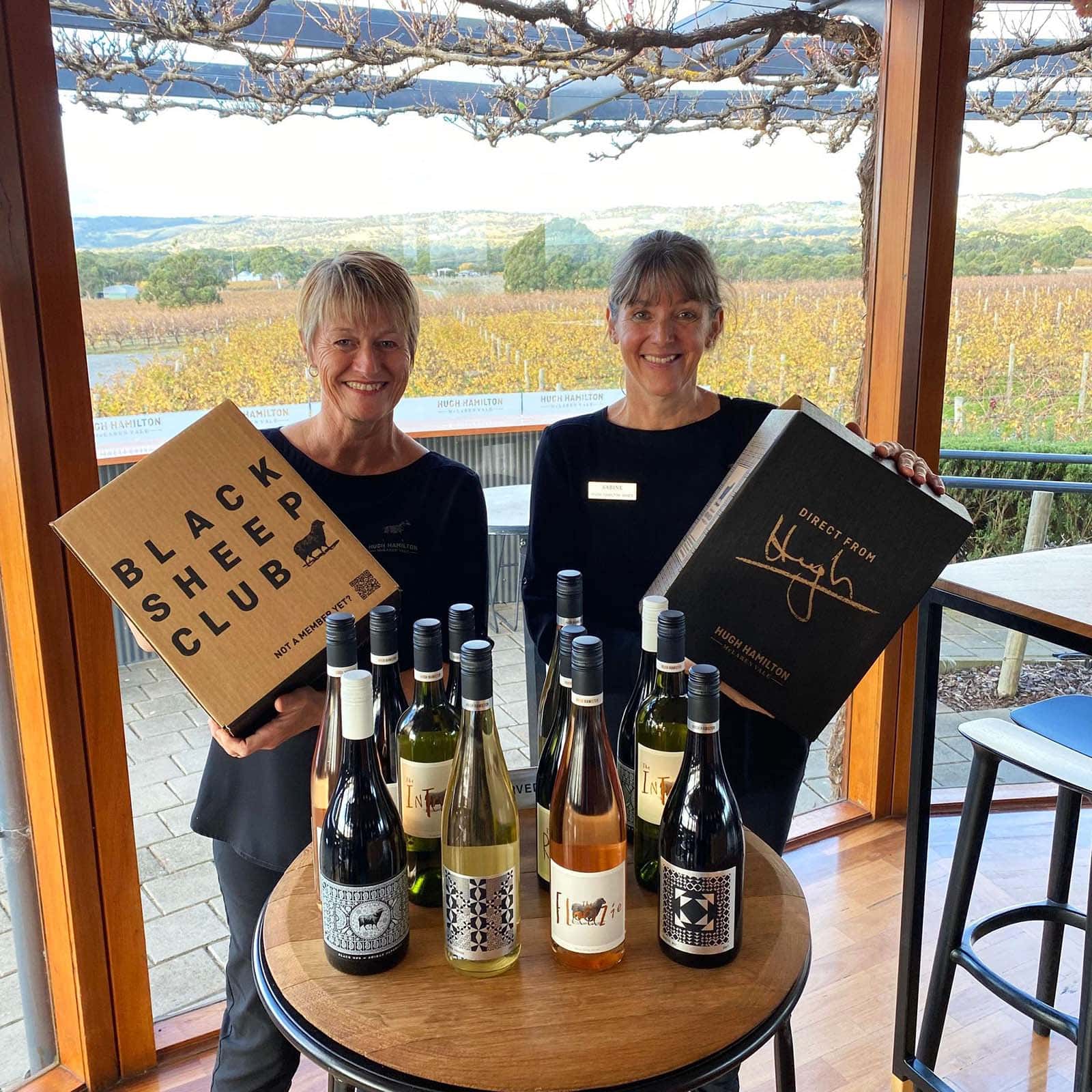 Two women holding boxes of Black Sheep Club wine, standing in front of a small table with 12 variations of Hugh Hamilton Wines. Behind them are rows of orange vines, and a view of a flat landscape. 