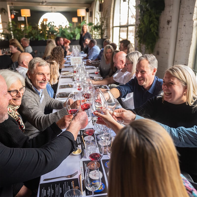 A long view of Black Sheep Club members sat along a table, the focus placed on members in the foreground, clinking their glasses and laughing. 