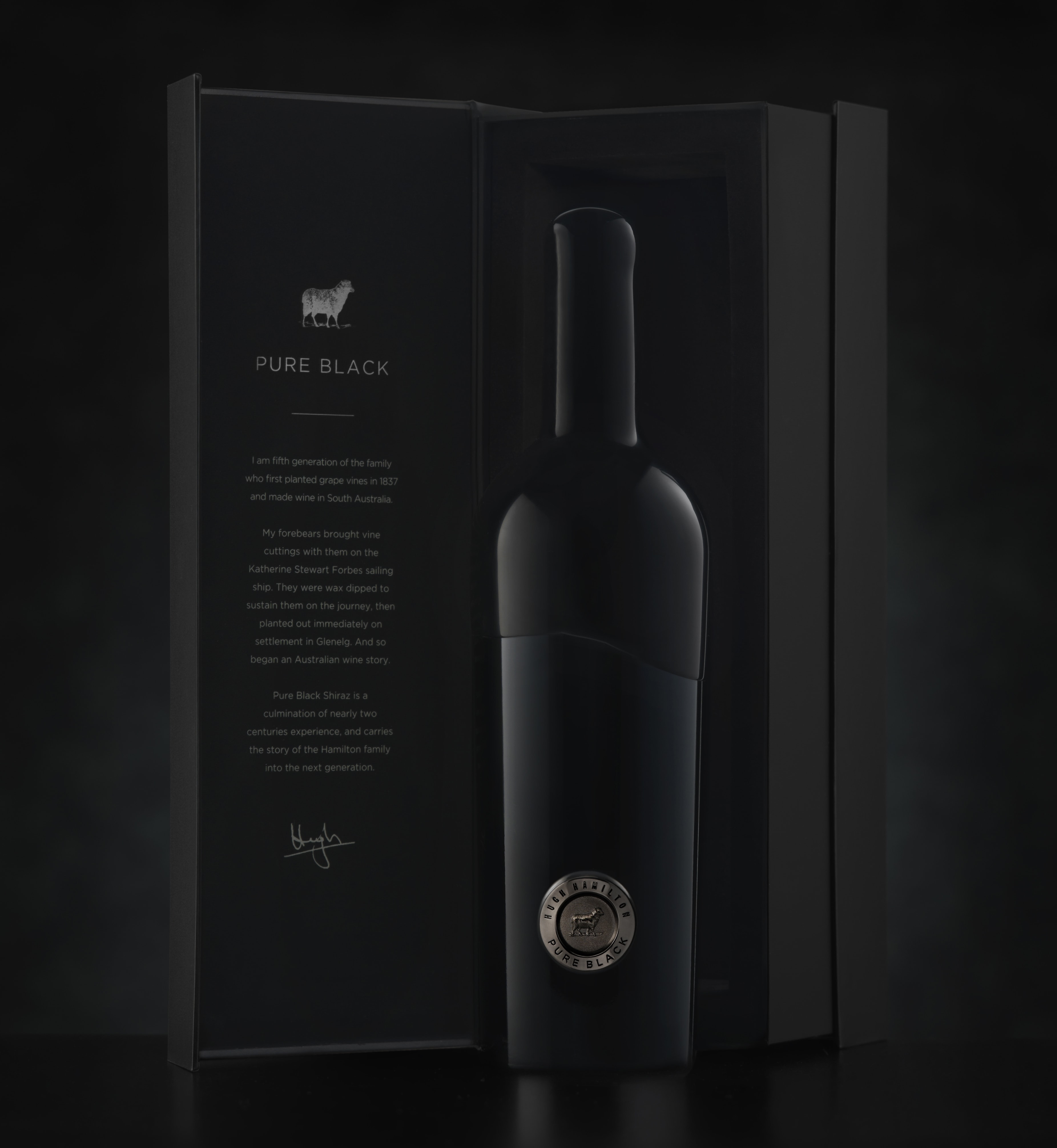 A photo of a black bottle of wine, posed in front of a black backdrop next to an open box for the wine, the Hugh Hamilton 'Pure Black' circular label placed near the bottom of the bottle, 