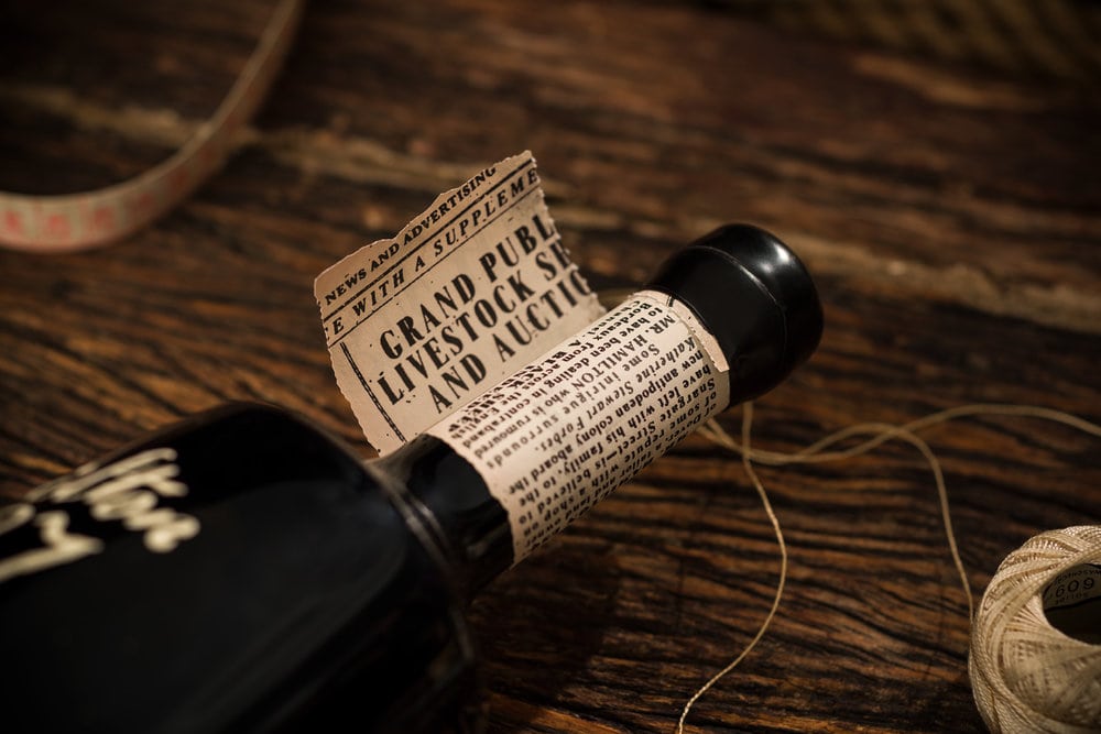 A close up of a wine bottles neck, a piece of news paper unfolding around it, twine and a measuring table placed alongside it on a wooden table. 