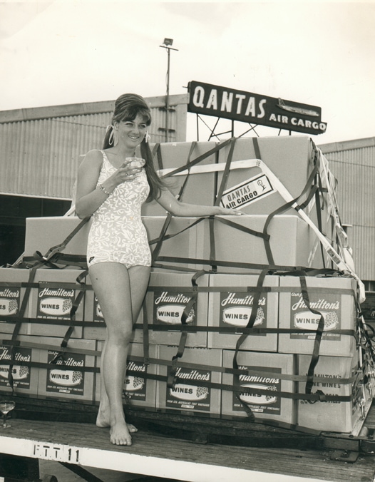 An old, black and white image of a young woman standing at an airstrip, posed in front of boxes of Hamilton Wines. 