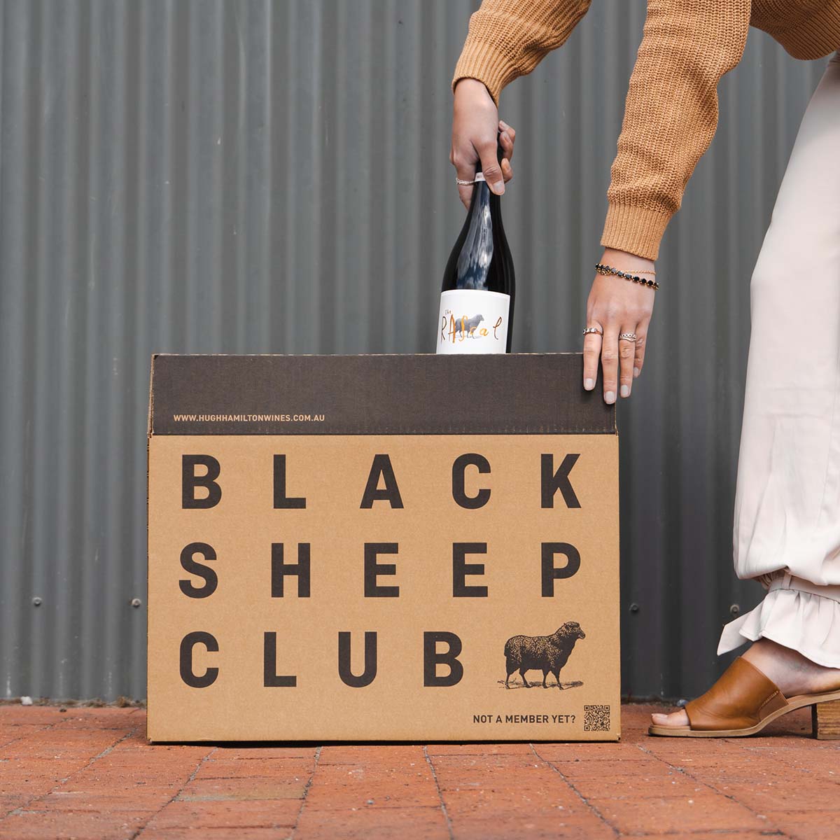 A Black Sheep Club box placed on brick flooring, a woman opening the box and pulling out a Hugh Hamilton Wines bottle, the label directed to the front. 