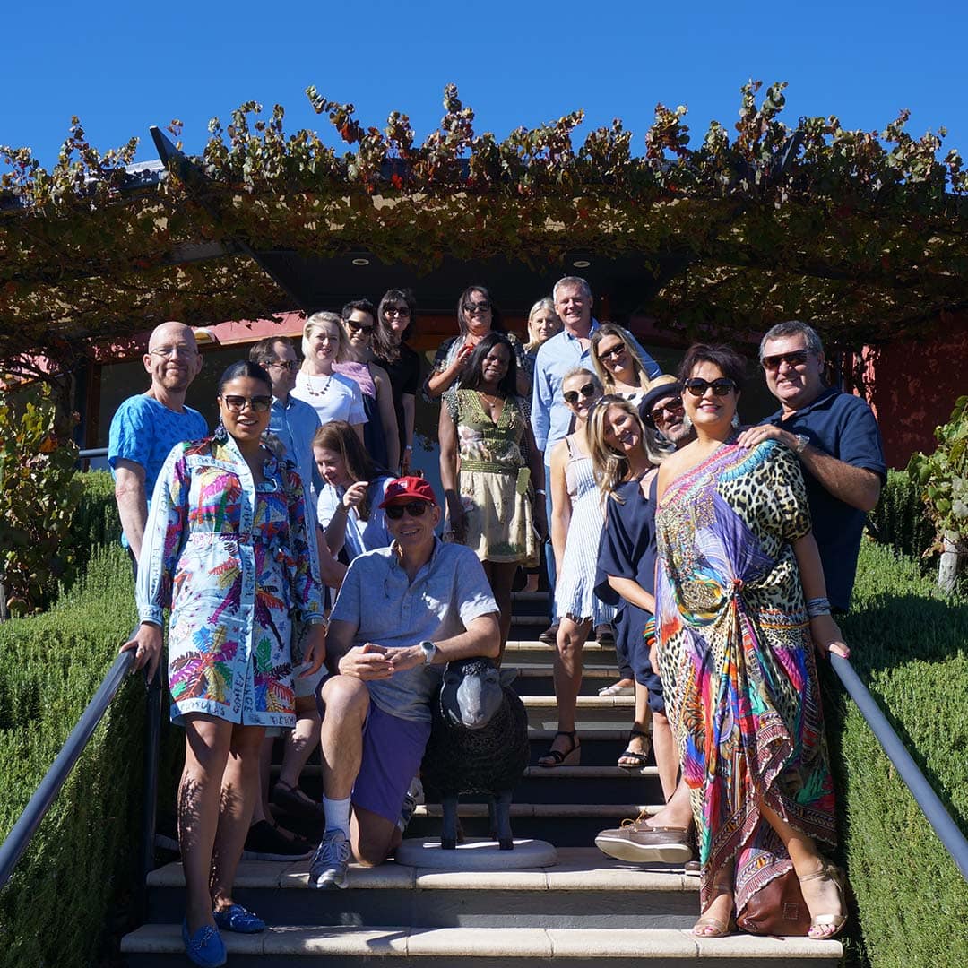 A collection of Hugh Hamilton Wines employees, smiling, posed on a stairway, with a covering of leaves, and blue skies above them. 