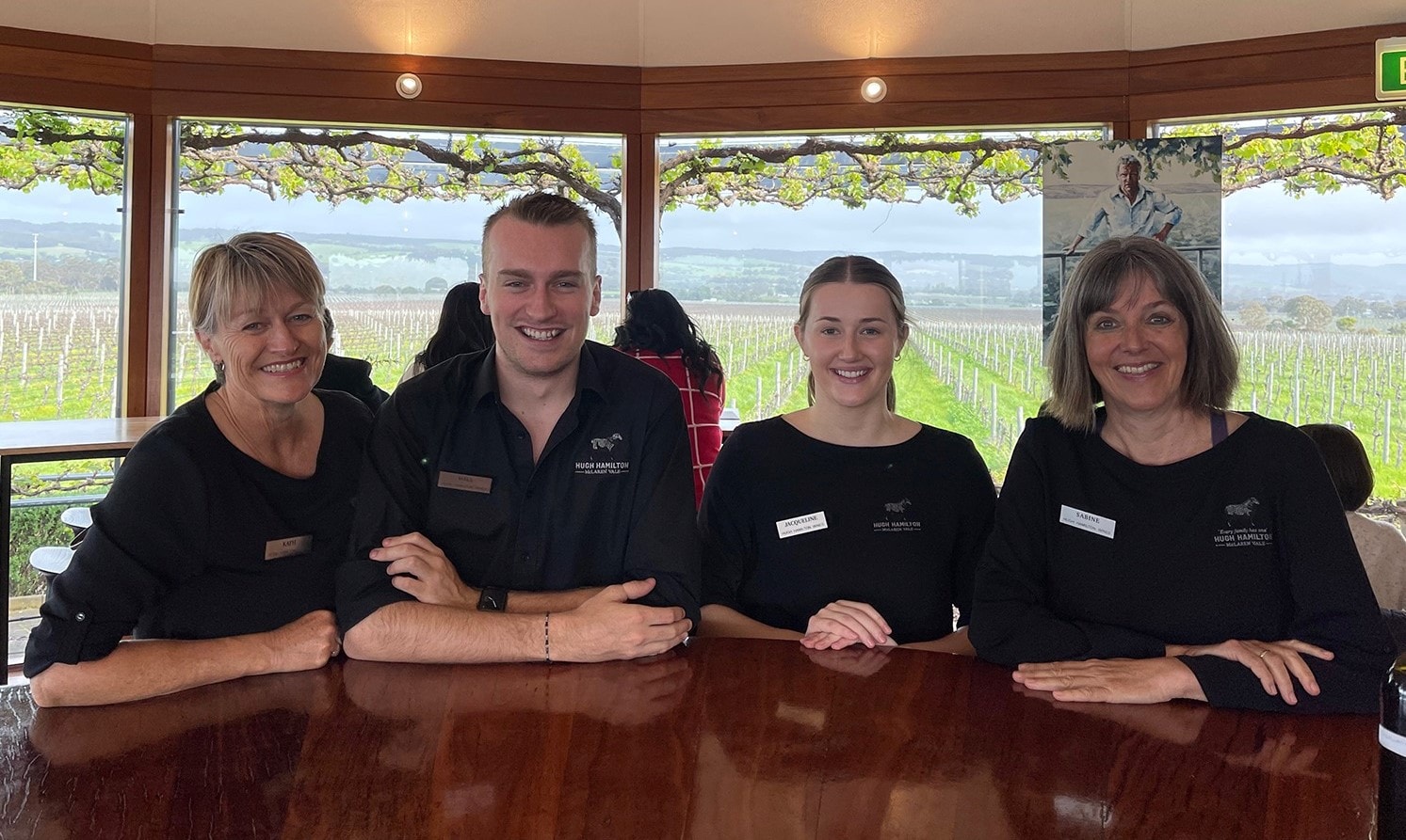 Four employees of Hugh Hamilton Wines, stood in a row along a table, smiling at the camera, with rows of vines seen outside behind them. 