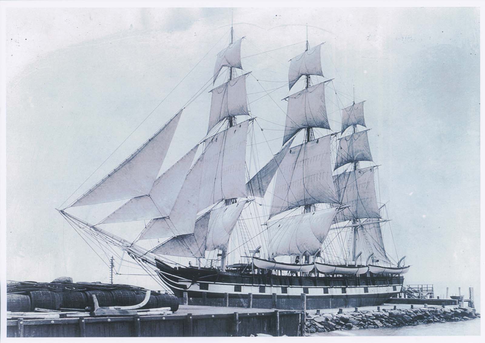 A black and white image of a ship with large, billowing sails, docked at a port. 