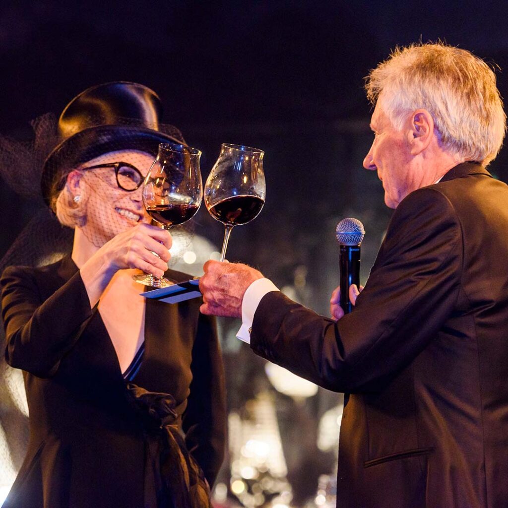 A man and woman smiling at each other, dressed formally, toasting their glasses together. 