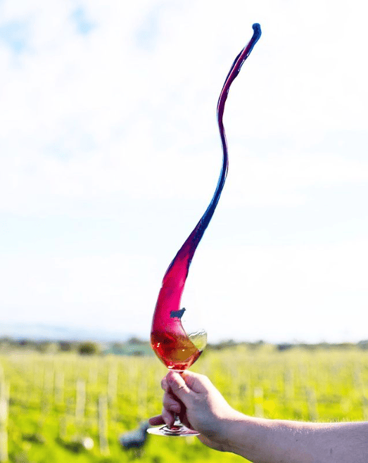 A man holding a glass of red wine, the wine shooting upwards in a swirling pattern, blurred skies and a vineyard in the background. 