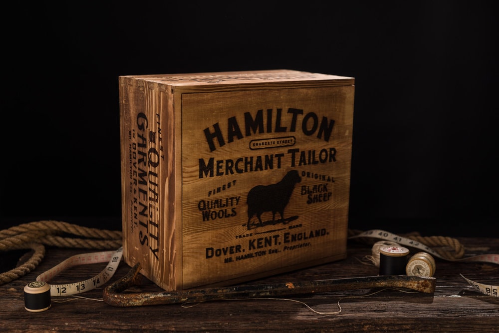 A wooden box, labeled Hamilton Merchant Tailor, placed on a wooden table with measuring tapes, rope, and thread - in front of a black backdrop. 